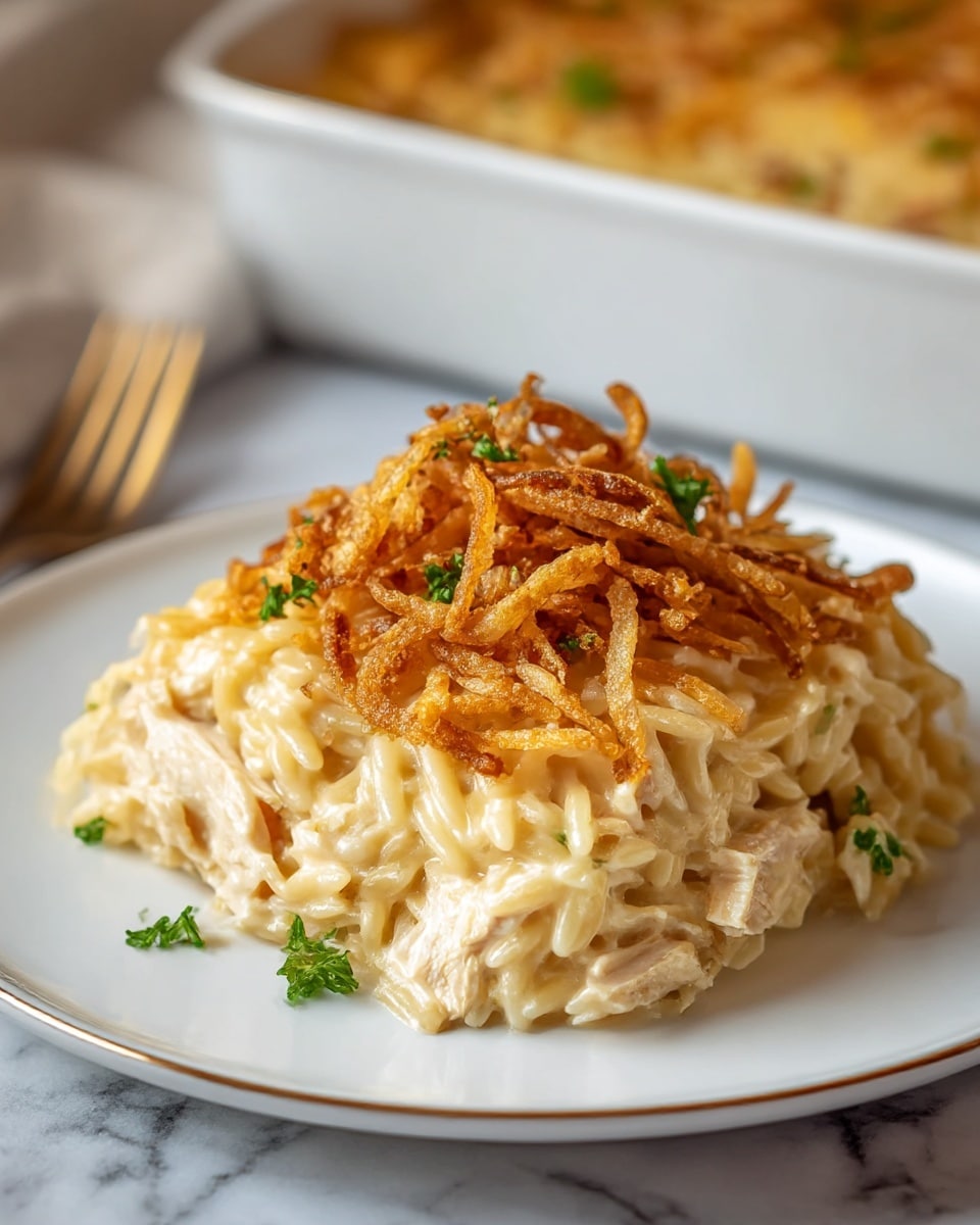 A white plate holds a creamy, three-layered dish of orzo pasta mixed with chunks of white chicken in a thick, pale yellow sauce. The top layer is a crispy pile of golden-brown fried onions scattered with small green parsley pieces. The creamy orzo forms the base, looking smooth and rich, with soft, tender chicken pieces embedded within. The crispy onions on top add texture and a darker golden contrast to the light tones beneath. The plate rests on a white marbled surface with a fork placed to the side, and a white baking dish with more of the dish is blurred in the background. Photo taken with an iphone --ar 4:5 --v 7