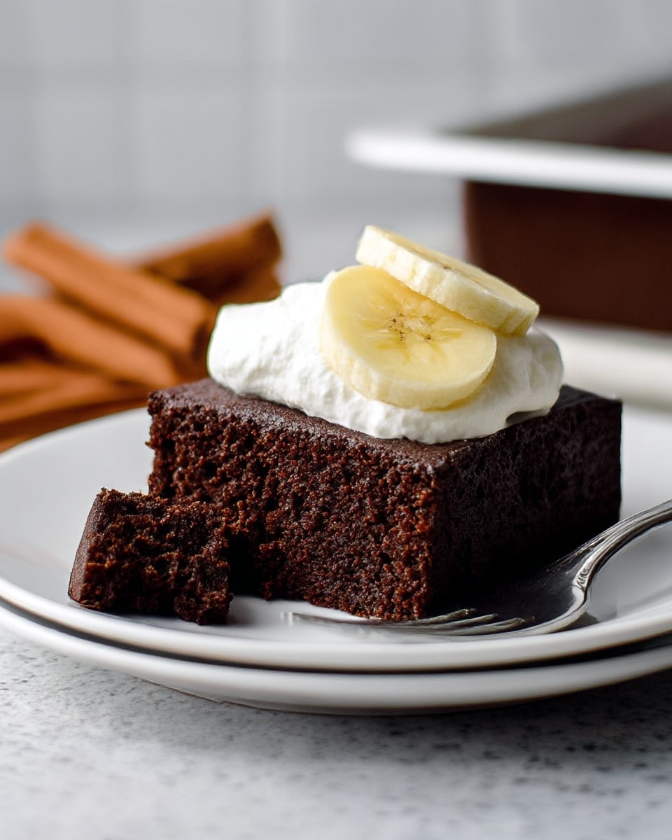 A thick, square piece of dark brown chocolate cake sits on a white plate with a silver fork resting beside it. The cake has a soft, moist texture shown by a small piece cut out on the left side. On top of the cake there is a layer of white whipped cream, and two light yellow banana slices are placed on the cream. The background shows a white marbled texture surface, with blurred cinnamon sticks lying near the plate. Photo taken with an iphone --ar 4:5 --v 7