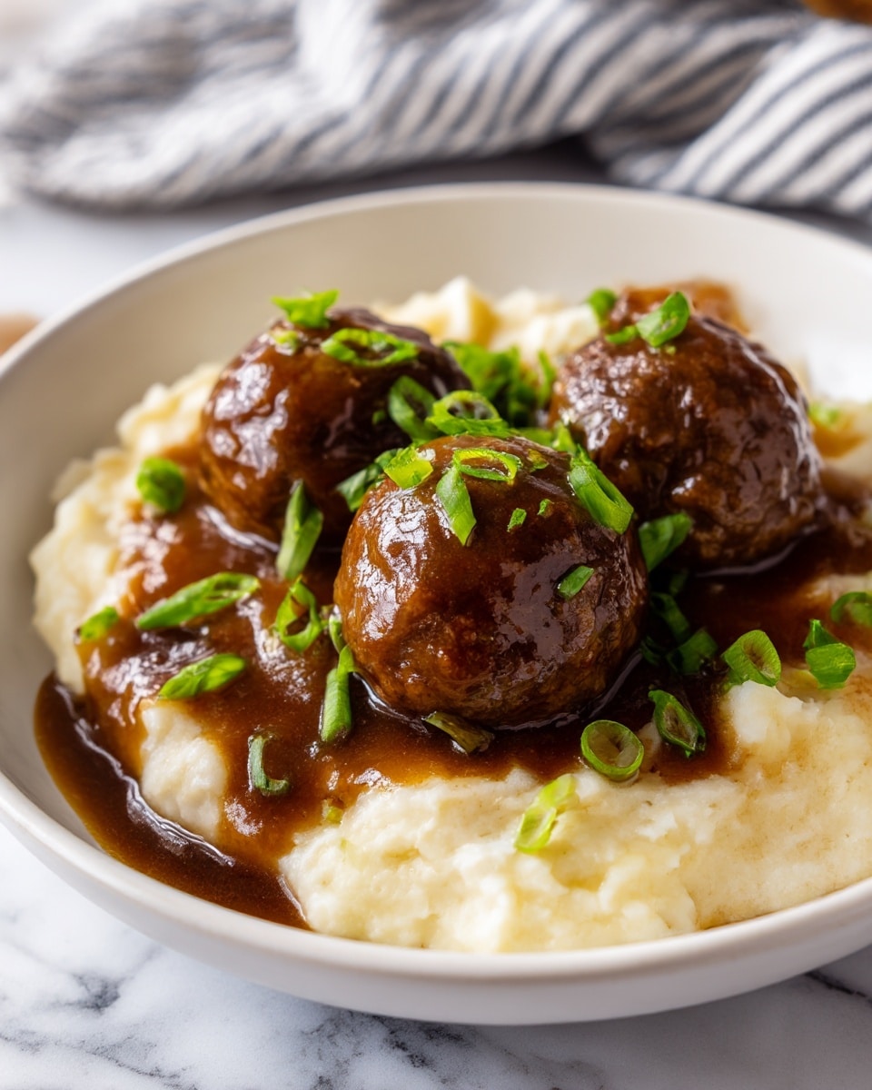 A close-up view of a dish with three visible dark brown meatballs covered in a shiny thick brown gravy on the top layer. These meatballs sit on a creamy, pale white mashed potato layer spread underneath. Small green pieces of chopped spring onions are sprinkled over the meatballs and gravy, adding a fresh green contrast. The food is served on a white plate, placed on a white marbled surface, with a blurred striped cloth in the background. Photo taken with an iphone --ar 4:5 --v 7