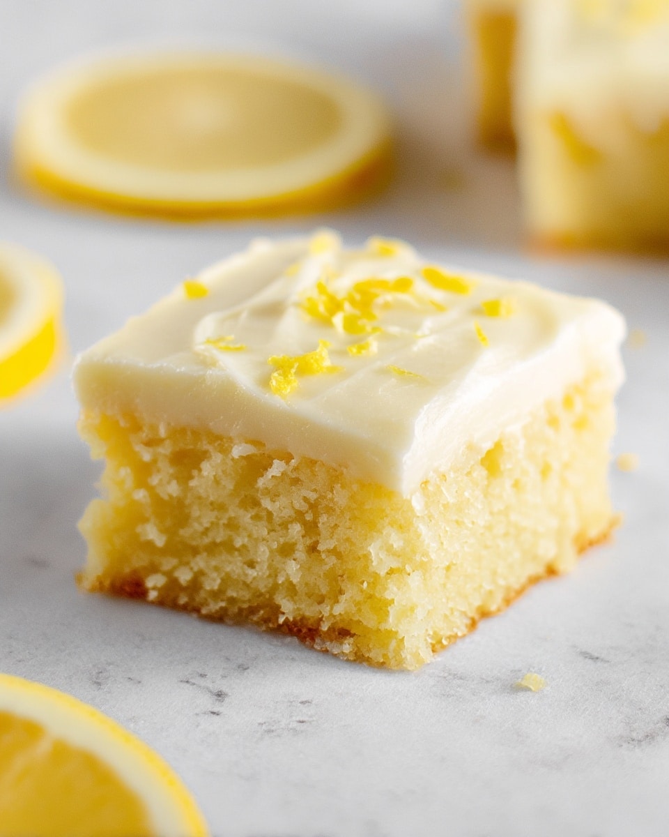 A close-up of a single square piece of lemon cake with two visible layers: the bottom layer is crumbly and light yellow with a soft texture, and the top layer is smooth, creamy, and pale yellow frosting with small lemon zest bits sprinkled on top. The cake is placed on a white marbled surface, with two thin lemon slices blurred in the background and one lemon slice closer to the bottom left corner. Photo taken with an iphone --ar 4:5 --v 7