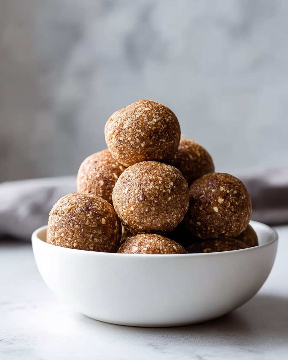 A white bowl filled with round, brown energy balls piled high, each ball having a coarse, crumbly texture with small bits visible on the surface, the balls showing shades of light to dark brown and stacked to create a small mound in the bowl. The background is a blurred white marbled texture, adding a soft, neutral backdrop that contrasts with the rich brown tones of the balls. photo taken with an iphone --ar 4:5 --v 7