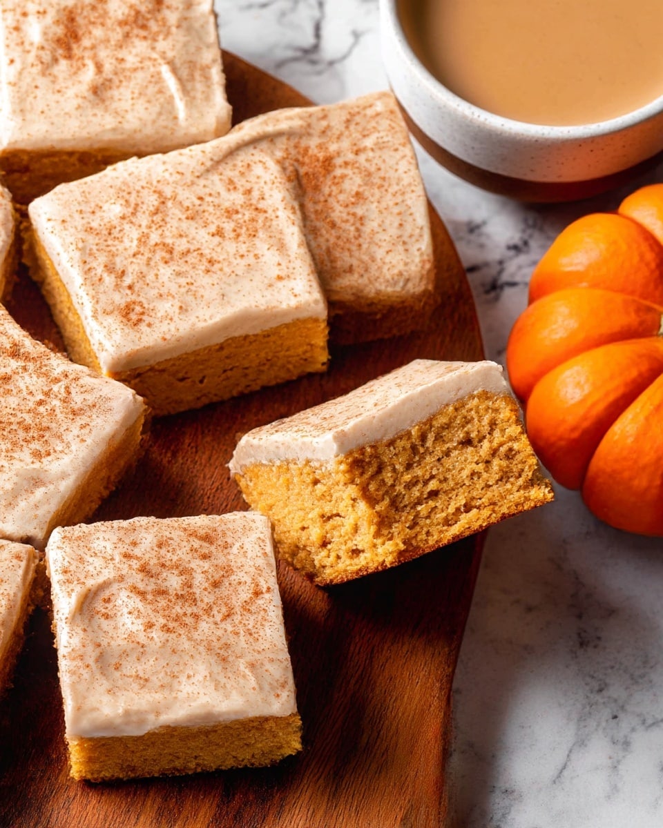 The image shows several square pieces of a moist, orange-brown cake topped with a thick layer of smooth, light tan frosting that is sprinkled evenly with fine cinnamon powder. One piece is lifted, revealing the soft and airy crumb texture inside. The cake pieces are placed on a wooden surface, with a small orange pumpkin on one side and a white bowl with a light brown creamy drink on the other. The background is a white marbled texture. photo taken with an iphone --ar 4:5 --v 7