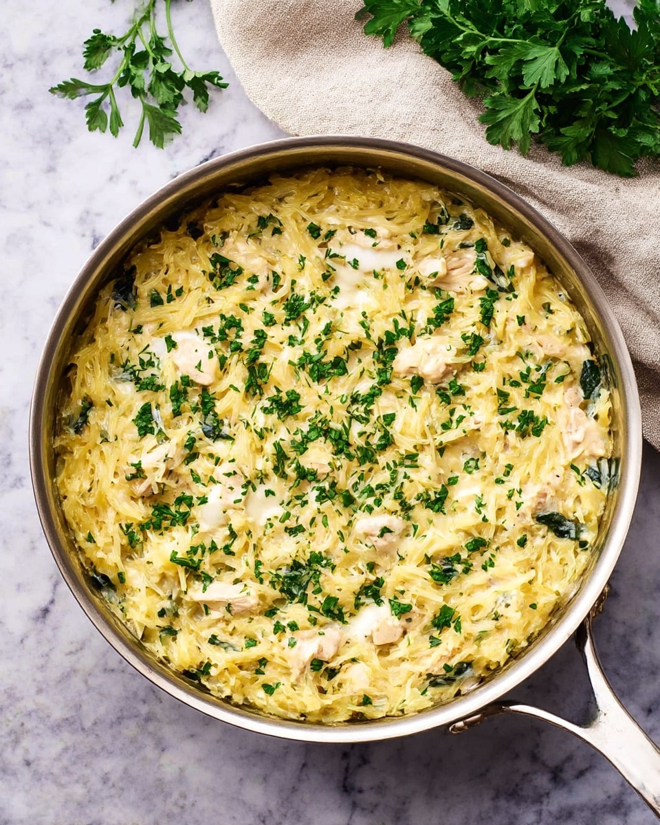 A large silver pan filled with a creamy dish made of shredded yellow spaghetti squash mixed with white melted cheese and pieces of cooked chicken. There are visible green spinach leaves scattered throughout the dish. The top is sprinkled with finely chopped green herbs, giving a fresh look. The pan sits on a white marbled surface next to a beige cloth and some fresh parsley leaves. photo taken with an iphone --ar 4:5 --v 7