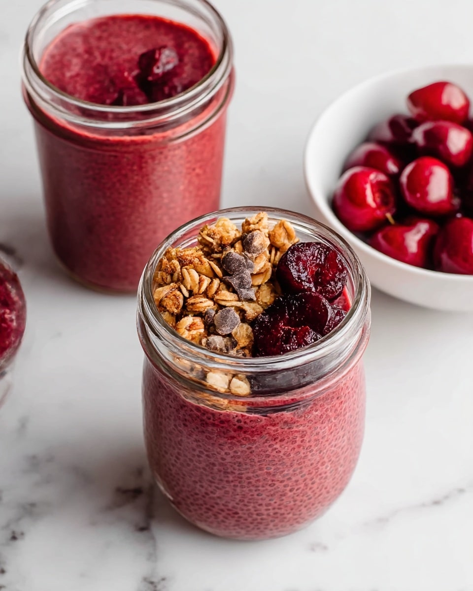 A clear glass jar filled with three distinct layers; the bottom and main layer is a thick, textured red chia pudding with visible chia seeds, filling most of the jar. On top, one side has a crunchy golden granola cluster layer, next to another small pile of dark brown chocolate chips, and the other side features a few dark red cherry pieces with a glossy, juicy texture. Above the jar, there is a second glass jar filled completely with the same red chia pudding, and to the right, a white bowl holds a few whole dark red cherries. The scene is set on a white marbled surface. photo taken with an iphone --ar 4:5 --v 7