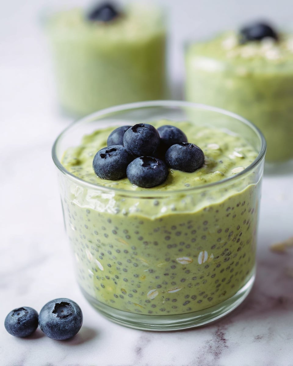 A clear glass cup filled with a creamy green mixture that contains small black seeds and oat flakes, showing one thick layer of this textured green pudding. On top, there is a small pile of five plump, deep blue blueberries standing out against the green base. Around the glass cup, a few more blueberries rest scattered on the white marbled surface, adding pops of dark blue color. In the blurred background, another similar glass with the same green mixture and a single blueberry on top is visible. The overall scene is bright and clean, with soft natural lighting. photo taken with an iphone --ar 4:5 --v 7
