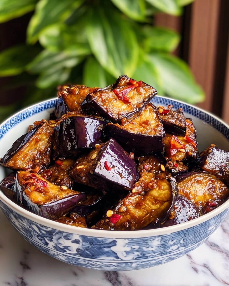 A white bowl with blue patterns is filled with many thick chunks of cooked eggplant that have a shiny, dark purple skin and a golden-brown inside, showing a slightly crispy texture. Each piece glistens with a sticky glaze that looks like a rich sauce with small bits of red chili and white seeds, adding a spicy touch. The bowl sits on a white marbled surface with a blurred green leafy background behind it. photo taken with an iphone --ar 4:5 --v 7