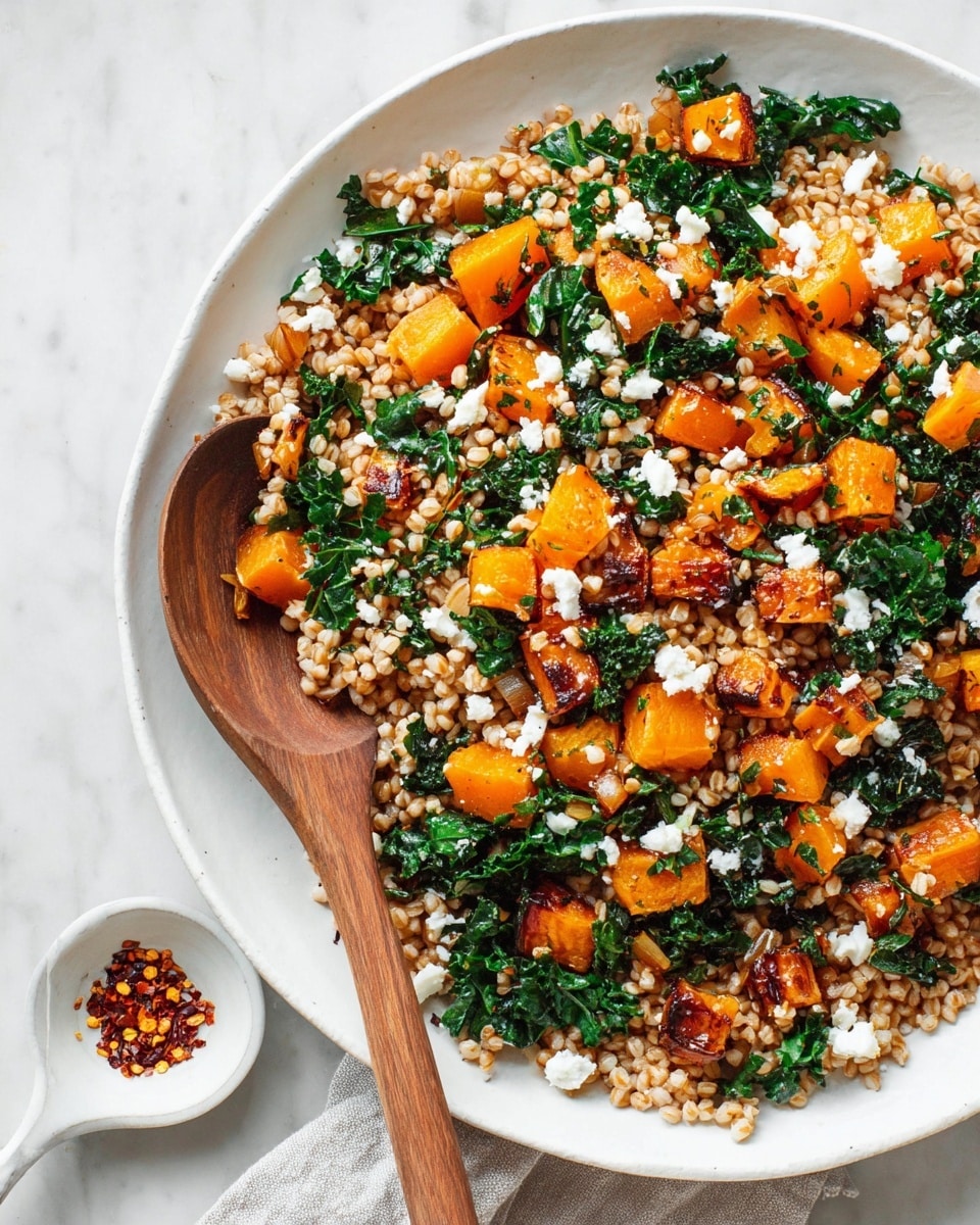 A large white plate with a mix of cooked wheat grains forming the base layer, topped with bright orange roasted butternut squash cubes and dark green wilted kale leaves scattered unevenly. Small white crumbles of feta cheese are sprinkled over the top. A wooden spoon is placed inside the dish on the left side, partially submerged in the food. The plate rests on a white marbled surface with a small white dish holding red chili flakes nearby. Photo taken with an iphone --ar 4:5 --v 7