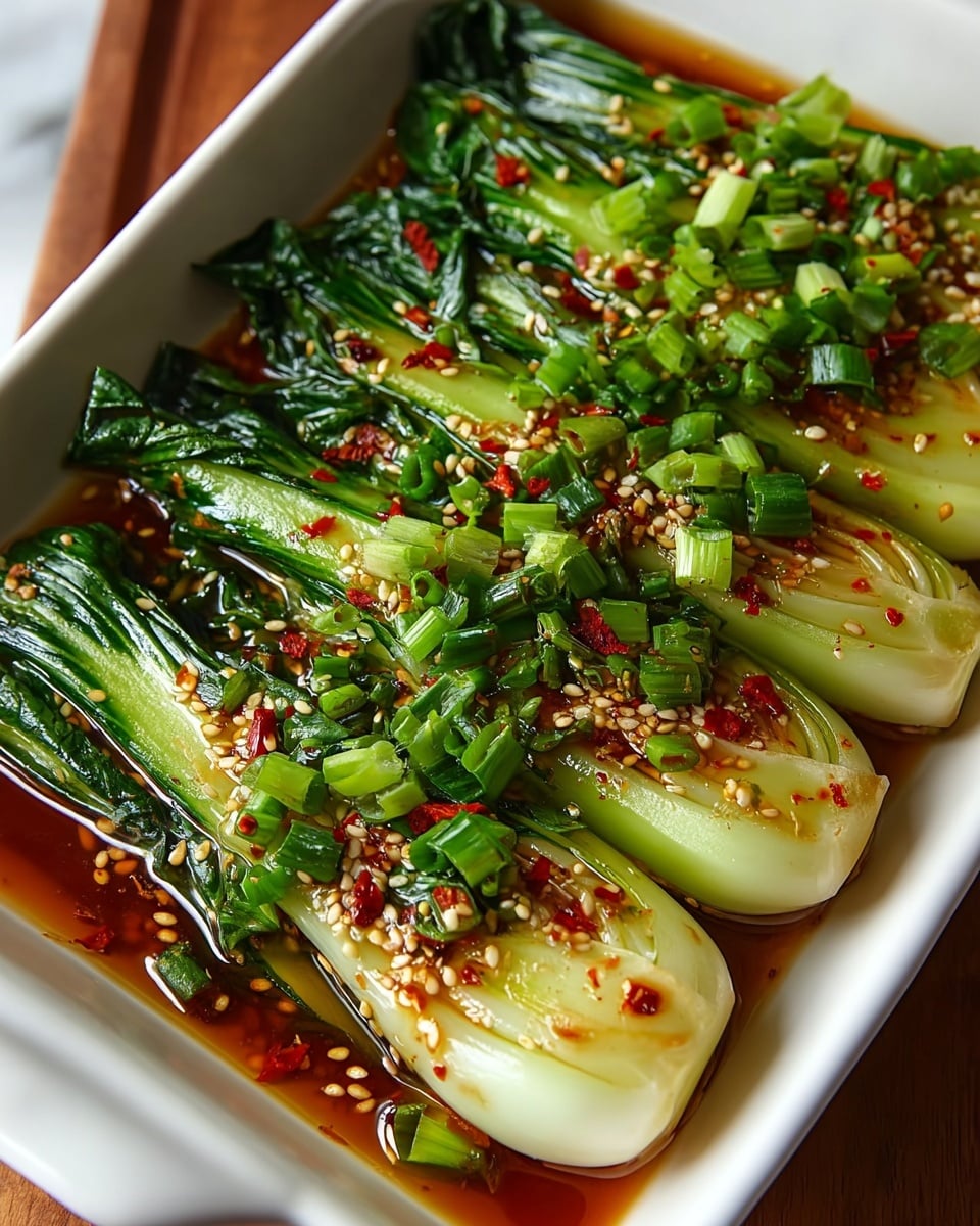 The image shows a white rectangular dish filled with five whole bok choy layered side by side. Each bok choy is pale green at the base and dark green at the leafy ends, softened by cooking. The vegetables are coated in a glossy brown sauce speckled with red chili flakes and small beige sesame seeds. On top, a generous layer of finely chopped bright green scallions adds texture and color contrast. The dish rests on a white marbled surface with a warm wooden edge slightly visible. Photo taken with an iphone --ar 4:5 --v 7
