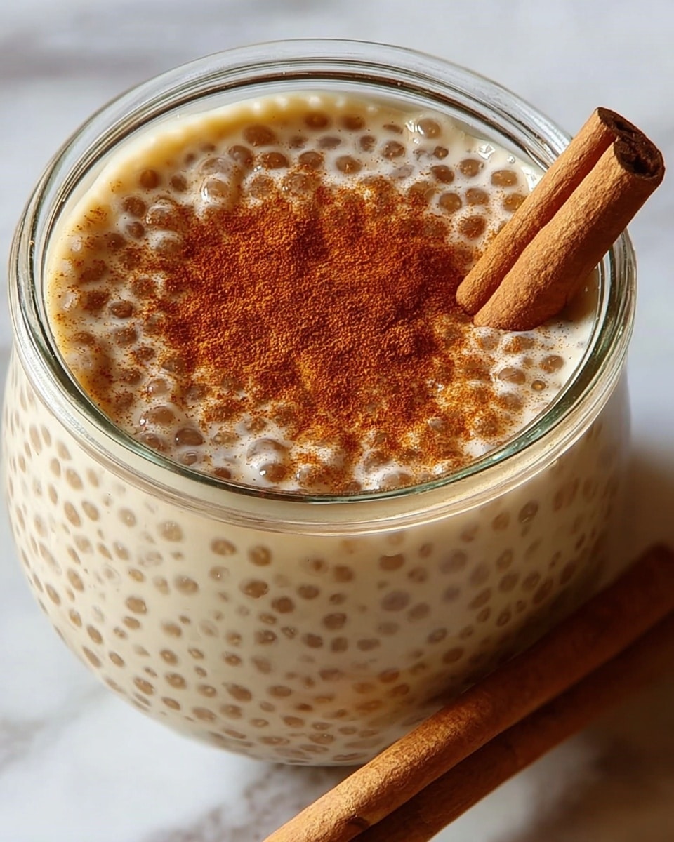 A close-up of a glass jar filled with a creamy beige pudding-like dessert with small translucent tapioca pearls evenly mixed throughout. On top, there is a light dusting of reddish-brown cinnamon powder. Two whole cinnamon sticks, light brown with a textured surface, rest diagonally at the edge of the jar. The jar is placed on a white marbled surface. photo taken with an iphone --ar 4:5 --v 7