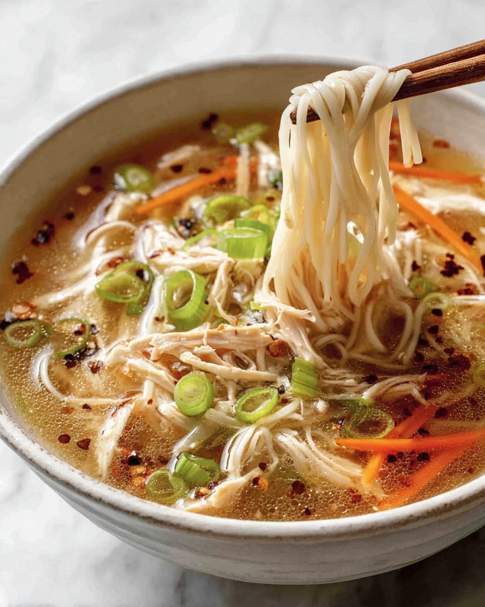 A white bowl filled with clear light brown broth, with soft thin white noodles lifted by wooden chopsticks, showing their smooth and slightly shiny texture. Inside the broth, there are shredded light brown chicken pieces scattered around, thin bright orange carrot sticks, and round green slices of spring onion floating on top. Small black chili flakes are sprinkled over the noodles and broth, adding contrast and texture. The bowl is placed on a white marbled surface. Photo taken with an iphone --ar 4:5 --v 7