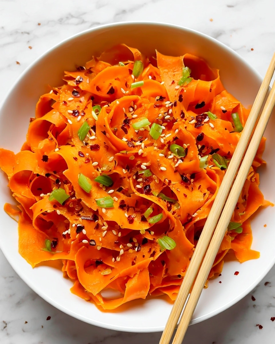 The dish shows a white bowl filled with thin, curly orange carrot ribbons that are lightly tossed. These carrot ribbons create a soft layered texture with a mix of smooth and folded strips. Scattered on top are small green onion pieces, adding pops of bright green color, and white sesame seeds spread evenly, giving a bit of texture contrast. There are also small red chili flakes lightly sprinkled around, adding a hint of red. A pair of light-colored wooden chopsticks is placed diagonally on the top right side, partly touching the carrot ribbons. The bowl sits on a white marbled surface. photo taken with an iphone --ar 4:5 --v 7