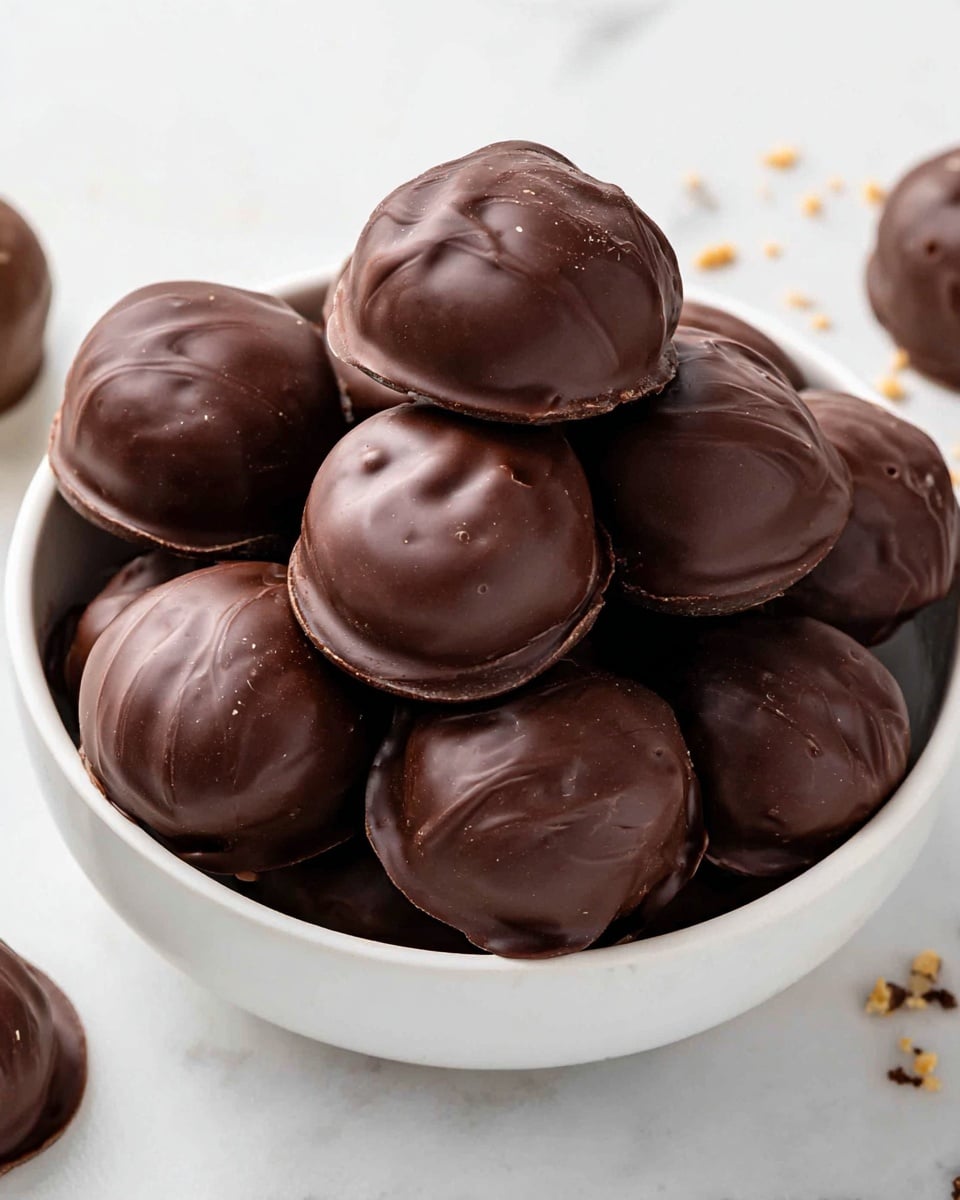 A white bowl filled with about a dozen round chocolate-covered treats. Each piece is fully coated in smooth, shiny dark brown chocolate with slight swirls and small textures on the surface, giving a rich look. The chocolates are closely packed, with some stacked on top, showing their round shape and thick chocolate shell. The bowl sits on a white marbled surface with a few crumbs and chocolate pieces scattered around. Photo taken with an iphone --ar 4:5 --v 7