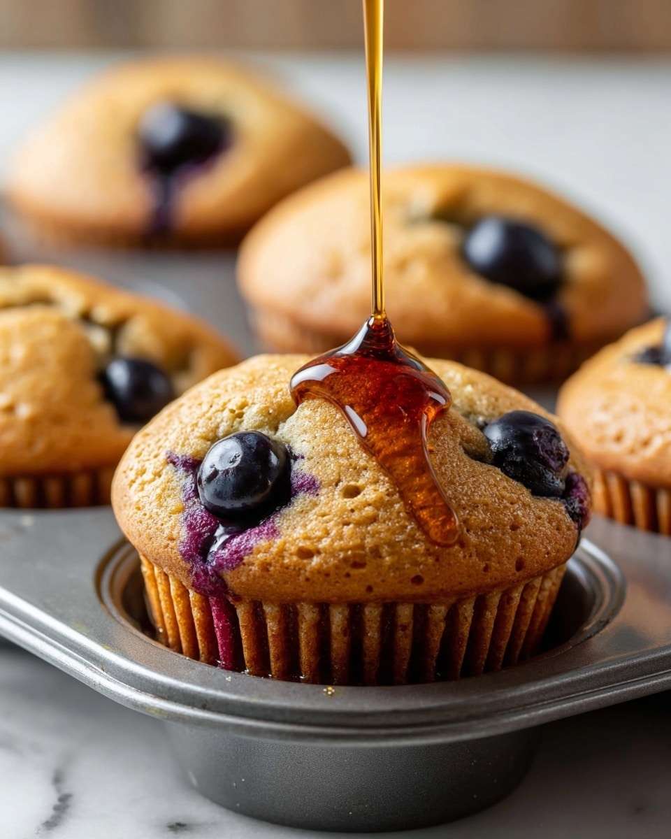 A close-up of a golden-brown blueberry muffin in a metal muffin tray with several whole blueberries baked into the top, some showing a deep purple color where the berries burst. A stream of shiny amber syrup is being poured over the muffin, creating a thick layer flowing down the side and pooling slightly at the base. The muffins are textured with small air bubbles on the surface, and the tray sits on a white marbled surface in soft natural light. photo taken with an iphone --ar 4:5 --v 7