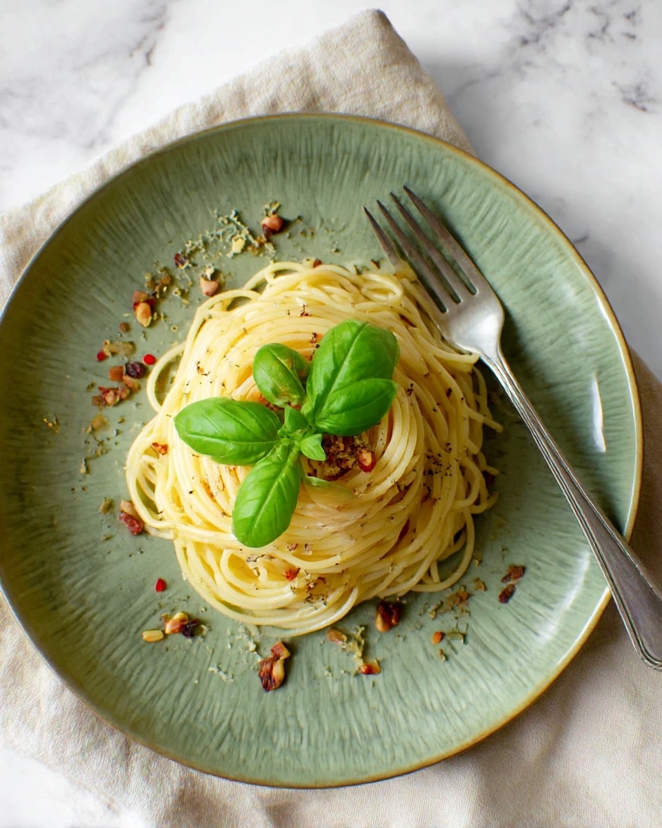 A plate with a textured green color holds a small mound of spaghetti pasta, twisted neatly in the center. The pasta is light yellow with a slight shine, mixed with small bits of herbs and red pepper flakes scattered throughout. On top of the spaghetti sits a fresh, bright green basil leaf cluster, adding a pop of fresh color. Around the pasta on the plate are small pieces of herbs and possibly bits of garlic or dried vegetables, adding texture. A silver fork rests on the edge of the plate. The plate is set on a beige cloth on a white marbled surface. Photo taken with an iphone --ar 4:5 --v 7