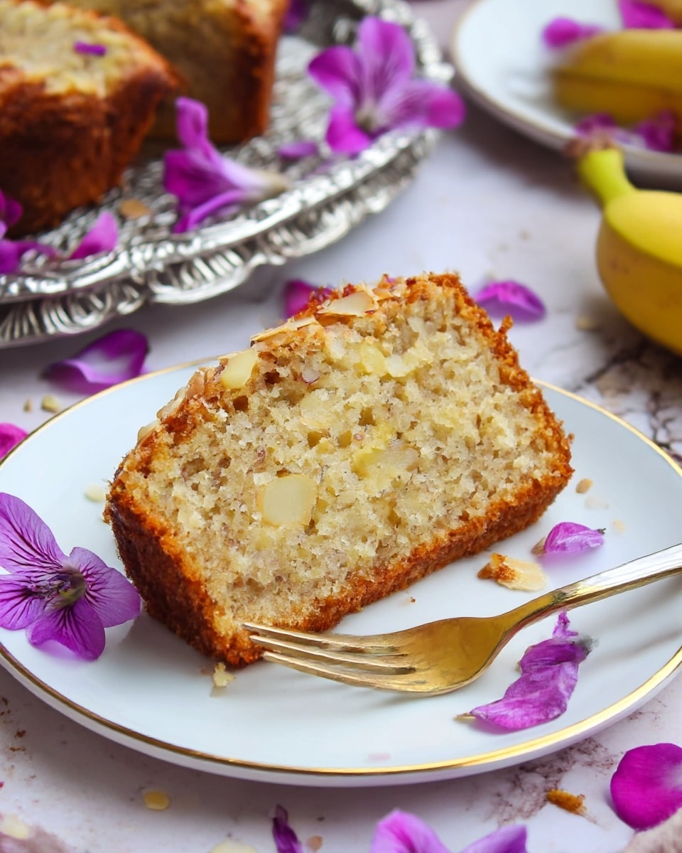 A thick slice of moist cake sits on a white plate with a thin gold rim, surrounded by soft purple flower petals scattered on and around the plate. The cake has a golden brown crust and a light yellow crumbly inside with visible bits of nuts or coconut. A small silver fork with a gold handle rests on the plate, touching the cake's right side. In the background, a silver decorative tray holds more cake pieces and purple flowers with a white marbled surface underneath. A partially visible banana is seen on the right side of the image. Photo taken with an iphone --ar 4:5 --v 7