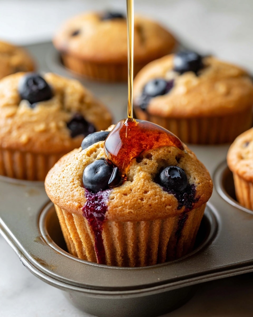 A close-up view of a golden brown blueberry muffin sitting in a gray metal muffin tray, with dark blue blueberries visible both on top and inside the muffin. A thick, dark amber syrup is being poured in a thin stream, flowing down the muffin's fluffy top and pooling slightly at the base in the tray. Behind it, other similar muffins with blueberries peek out softly blurred. The background features a white marbled texture. Photo taken with an iphone --ar 4:5 --v 7