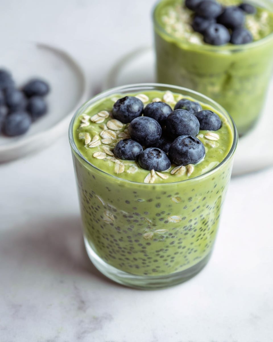 A clear glass cup filled with a layered green mix that has a creamy texture with visible oats and small black chia seeds spread throughout. The top layer is slightly thicker with oats more visible, and it is garnished with a cluster of fresh dark blue blueberries sitting on top. Surrounding the main cup, part of another similar cup is visible, also filled with the green oat mixture and topped with a blueberry. In the background on a white marbled surface, there is a white plate with a few scattered blueberries. The image uses soft lighting and a close-up view to highlight the fresh and healthy ingredients. photo taken with an iphone --ar 4:5 --v 7
