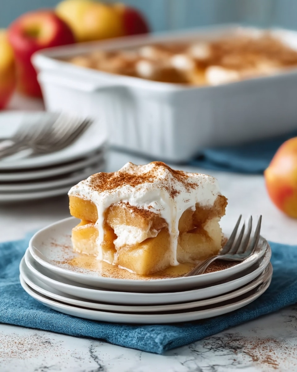 A dessert with two main layers sits on a stack of three white plates on a blue cloth over a white marbled surface. The bottom layer is a thick apple filling with soft, golden-brown apple slices mixed with a cinnamon-spiced sauce. Above it is a creamy white whipped topping, dusted with ground cinnamon, slightly melting and dripping down the sides. A silver fork is placed beside the dessert on the top plate. In the blurred background, there is a white baking dish filled with more of the apple dessert, along with red and yellow apples and a stack of white plates with forks. Photo taken with an iphone --ar 4:5 --v 7