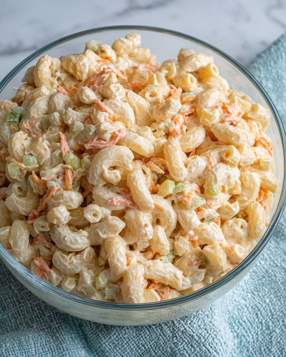 A clear glass bowl filled with a creamy macaroni salad, showing about one thick layer of elbow macaroni pasta mixed with small pieces of shredded carrot and green celery. The dish has a light, creamy dressing coating the pasta, giving it a pale, slightly yellow and off-white color with visible bits of orange and green. The bowl sits on a white marbled surface with a textured light blue cloth partially visible underneath. photo taken with an iphone --ar 4:5 --v 7