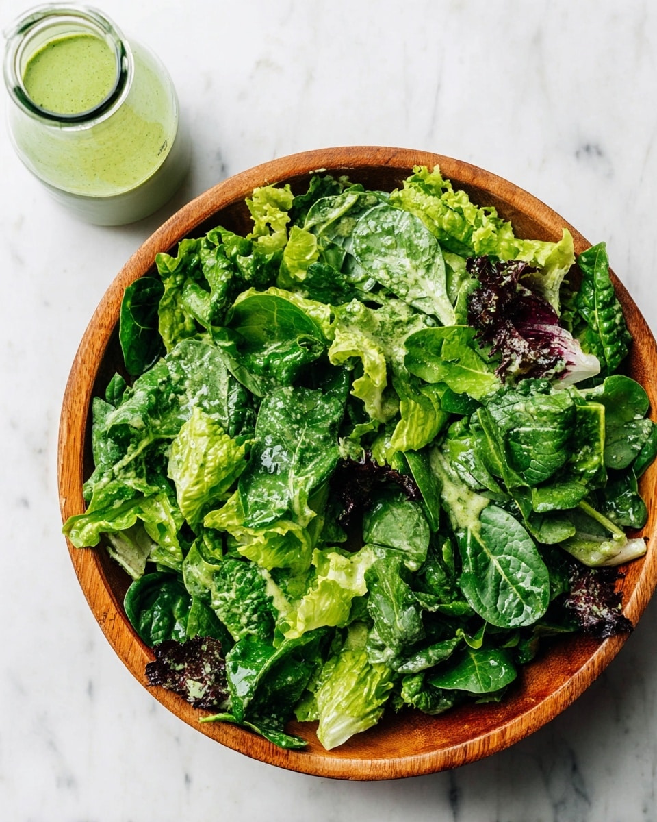 A close-up view of a wooden bowl filled with fresh mixed green salad leaves, including spinach, romaine, and some dark leafy greens, all lightly coated with a green dressing. The leaves are layered unevenly, showing a mix of different shapes and textures, with some curly edges and smooth flat leaves. To the top left of the bowl, there is an open bottle of green dressing with some dressing around the neck, all placed on a white marbled surface. photo taken with an iphone --ar 4:5 --v 7