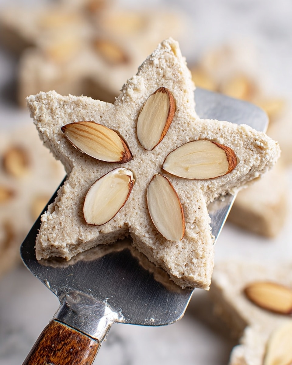 A star-shaped cookie with a pale beige color and a rough, slightly cracked surface is shown on a metal spatula with a wooden handle. The cookie has five whole almond slices embedded symmetrically on top, each with a light cream center and thin, dark brown edges. The background is a soft blur of more similarly shaped cookies resting on a white marbled surface. photo taken with an iphone --ar 4:5 --v 7