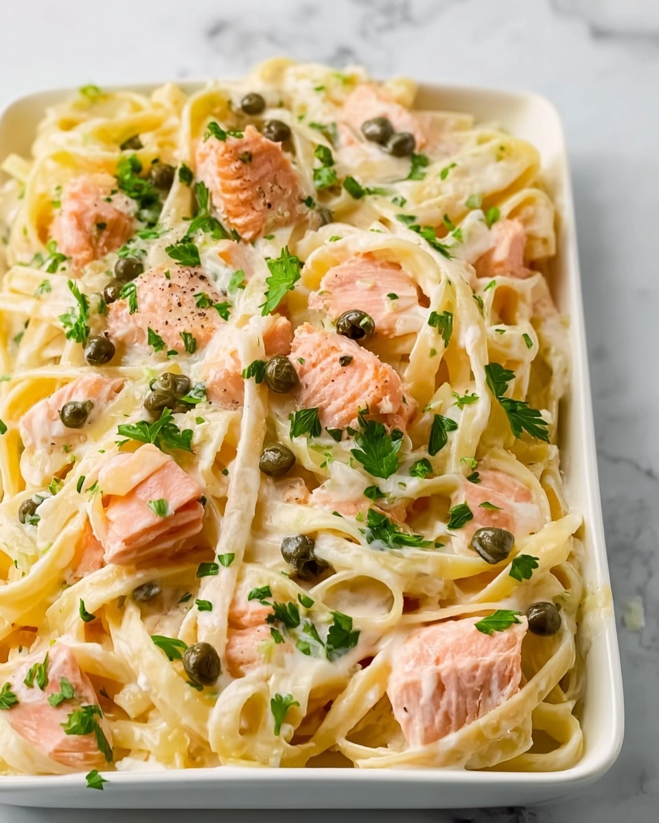 The image shows a white rectangular dish filled with fettuccine pasta coated in creamy white sauce. Scattered throughout the pasta are chunks of pink cooked salmon, small green capers, and sprinkled with fresh chopped green parsley. The creamy sauce covers the pasta evenly, giving it a smooth texture, and the salmon pieces are distributed on top and within the noodles. The white marbled surface beneath the dish adds a clean and bright look to the scene. photo taken with an iphone --ar 4:5 --v 7