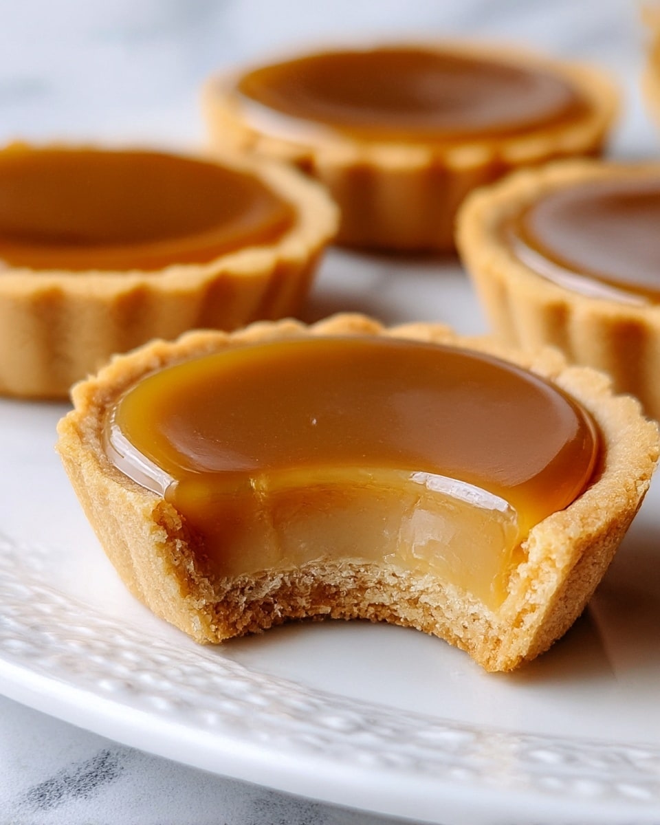 A close-up view of a small tart with two main layers: a thick, smooth, shiny caramel layer in a rich golden brown color on top, gently curved and glossy, and a light tan, crumbly tart crust at the bottom and sides, showing a bite taken out of the front tart revealing its soft, slightly chewy texture. Three similar tarts are partly visible, sitting on a white plate with a detailed raised pattern, all set against a white marbled texture. photo taken with an iphone --ar 4:5 --v 7