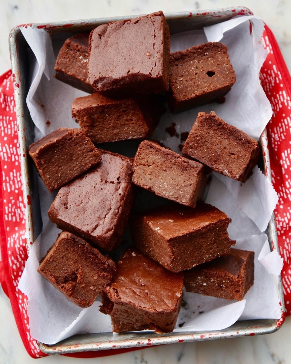 A metal tray lined with white parchment paper holds a group of thick, square brownies with a rough, slightly cracked surface. The brownies vary in shade from medium to dark brown, showing a dense and fudgy texture with a few small holes and crumbs around them. Some brownies are stacked in pairs while others lie flat, all arranged close together with irregular edges. The tray has red and white patterned sides, and it sits on a white marbled surface. Photo taken with an iphone --ar 4:5 --v 7