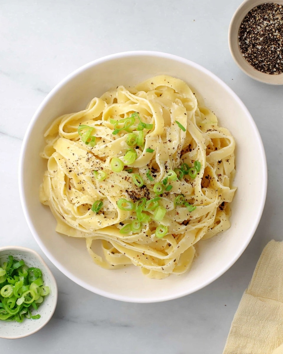 A white bowl filled with creamy, light yellow fettuccine pasta arranged in loose, twisted layers. The top is sprinkled with small green slices of scallion and a dusting of black pepper, adding specks of color and texture. The bowl sits on a white marbled surface, with small bowls containing ground black pepper and chopped green scallions nearby, completing the simple, fresh presentation. photo taken with an iphone --ar 4:5 --v 7