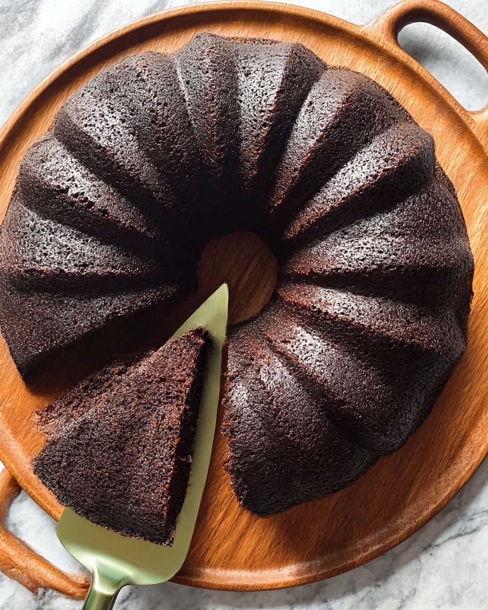 A dark chocolate bundt cake with a smooth, slightly glossy texture is shown on a round wooden tray with handles. The cake has a rich, almost black-brown color with finely textured surface and deep ridges running from the center to the edges. One slice is cut and being lifted with a cake server that has a green handle on a white marbled surface. The inside of the cake looks moist and dense, matching the dark outer layer. photo taken with an iphone --ar 4:5 --v 7