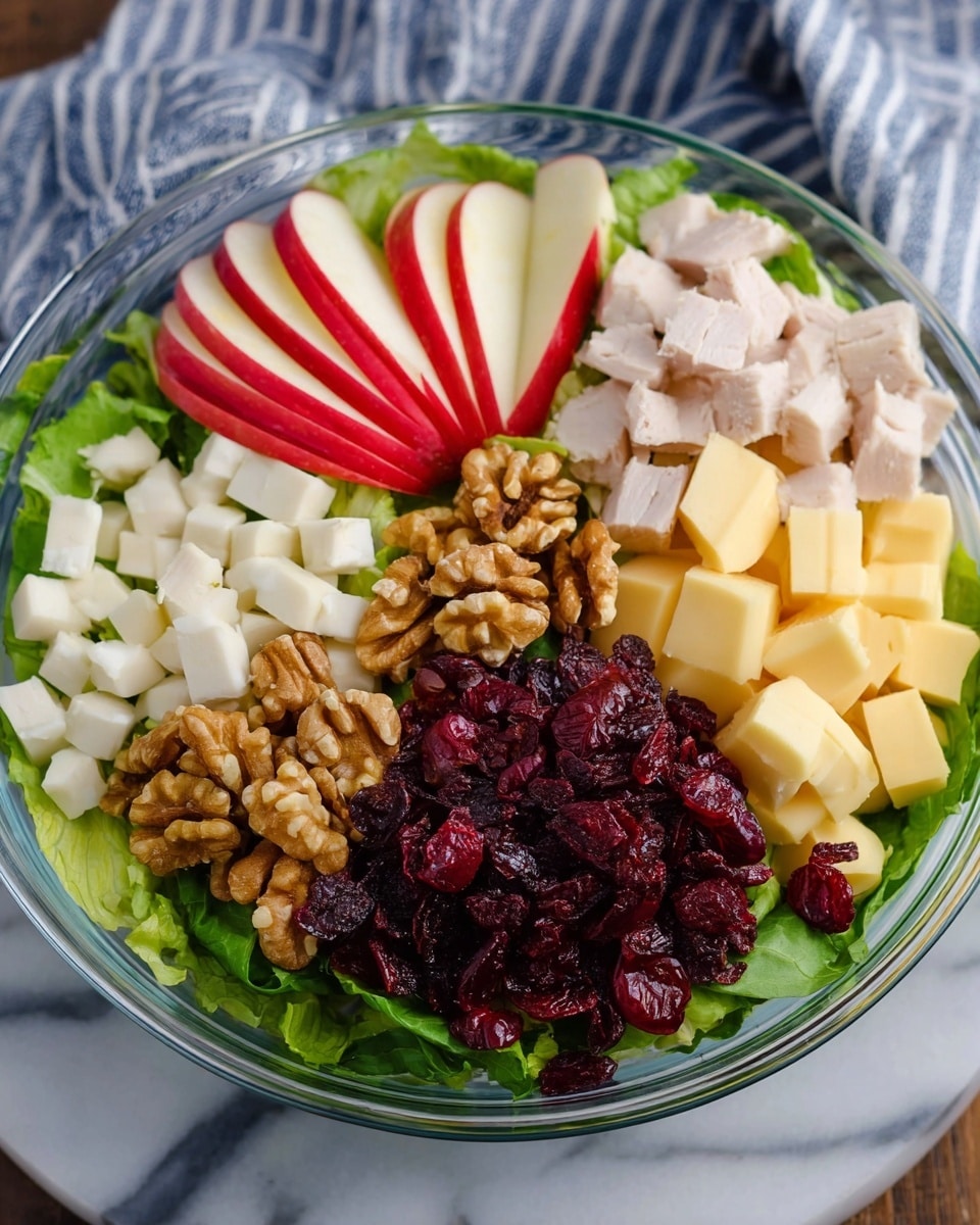 A clear glass bowl filled with green leafy lettuce at the bottom forms the base layer. On top, arranged in sections, are bright red apple slices with smooth skin and white inside on the left side, small white cubes of cooked turkey meat positioned on the top right, light yellow cheese cubes on the far right, a cluster of golden-brown walnut halves placed near the bottom left, and a pile of dark red dried cranberries near the bottom right over the greens. The bowl sits on a white marbled surface with a blue and white striped cloth partially visible in the background. Photo taken with an iphone --ar 4:5 --v 7