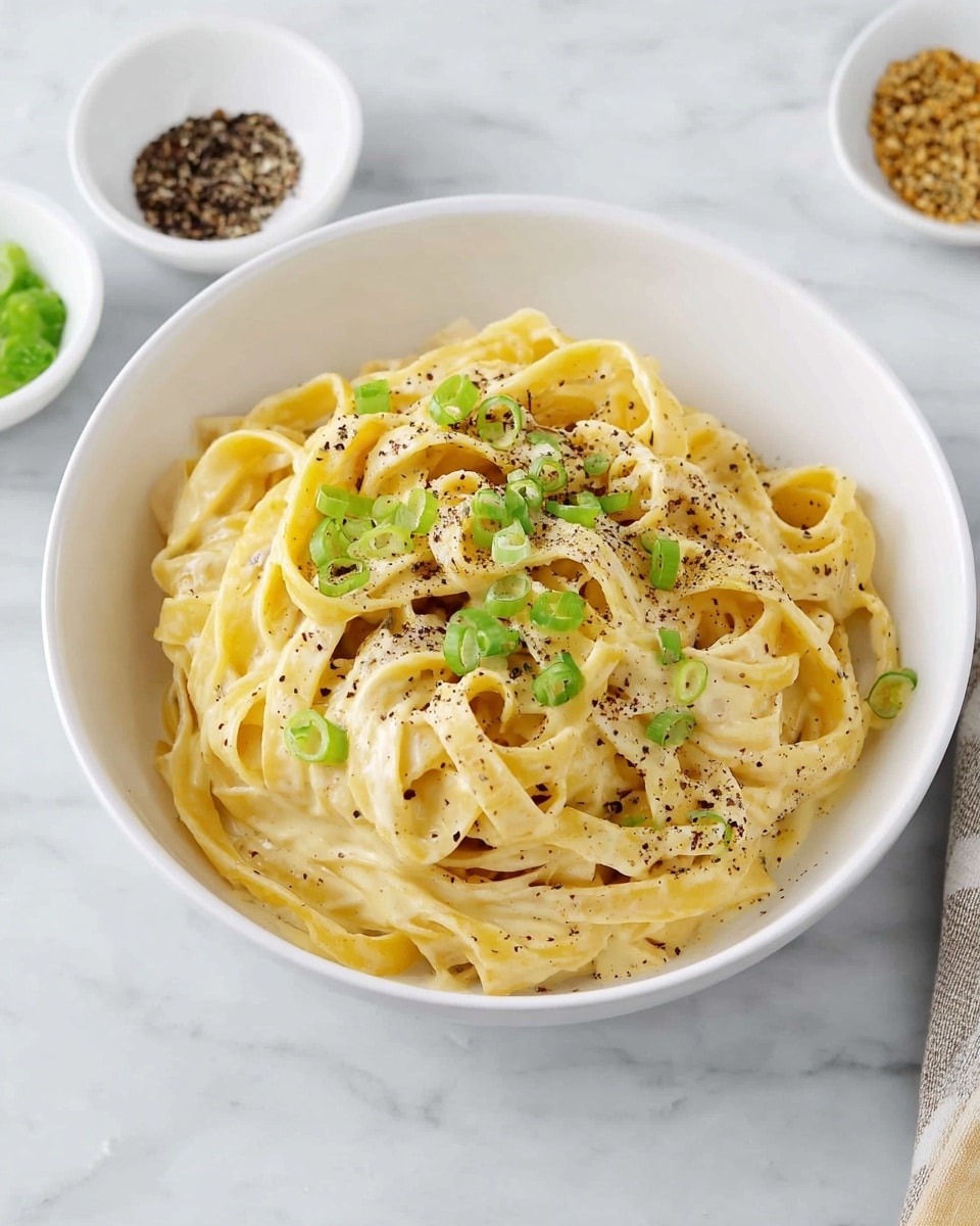 A white bowl filled with creamy fettuccine pasta, coated in a smooth, light yellow sauce. The noodles are thick and slightly curled, piled high in the center of the bowl. Thin slices of green onions are scattered on top, adding small touches of green, and a light sprinkle of black pepper dots the surface, giving a textured look. The bowl sits on a white marbled surface with small white dishes containing pepper and green onions nearby. photo taken with an iphone --ar 4:5 --v 7