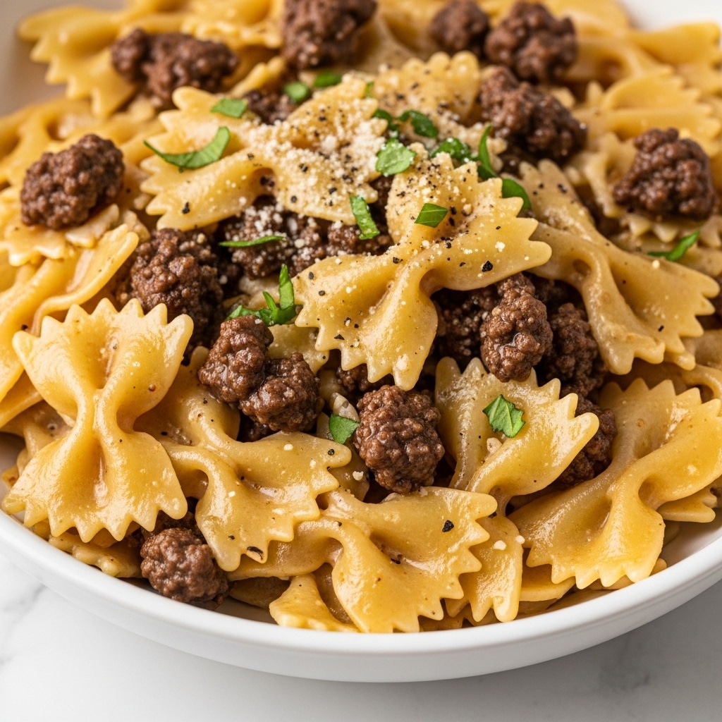 A close-up view of a white bowl filled with one layer of bowtie pasta mixed with browned ground beef. The pasta is a light golden-yellow color, slightly glossy from sauce, with edges that look soft and textured. The ground beef pieces are dark brown and crumbly, scattered evenly among the pasta. Small green herb pieces and a light sprinkle of grated cheese and black pepper are visible on top, adding a bit of color contrast. The bowl sits on a white marbled surface. Photo taken with an iphone --ar 4:5 --v 7