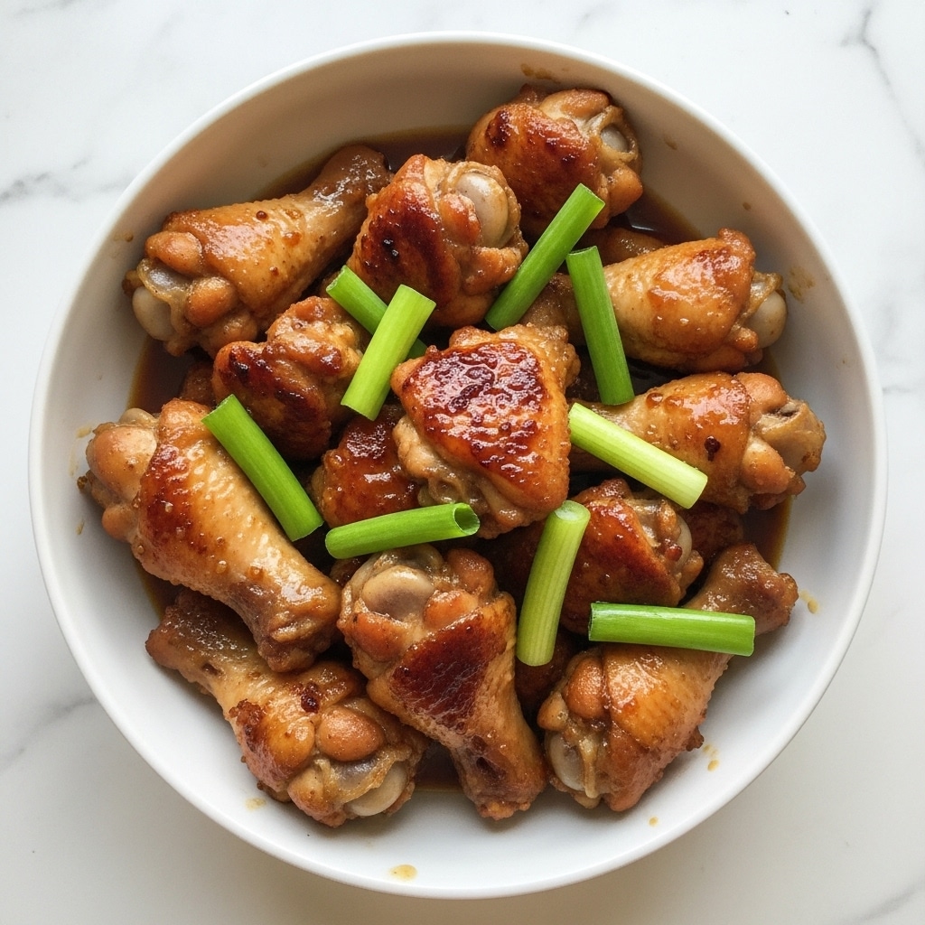 A white bowl filled with browned, cooked chicken pieces, varying in sizes and shapes like wings and drumettes, covered in a glossy sauce that looks thick and rich. Among the chicken pieces are scattered bright green whole spring onions, adding color contrast. The bowl is placed on a white marbled surface. photo taken with an iphone --ar 4:5 --v 7
