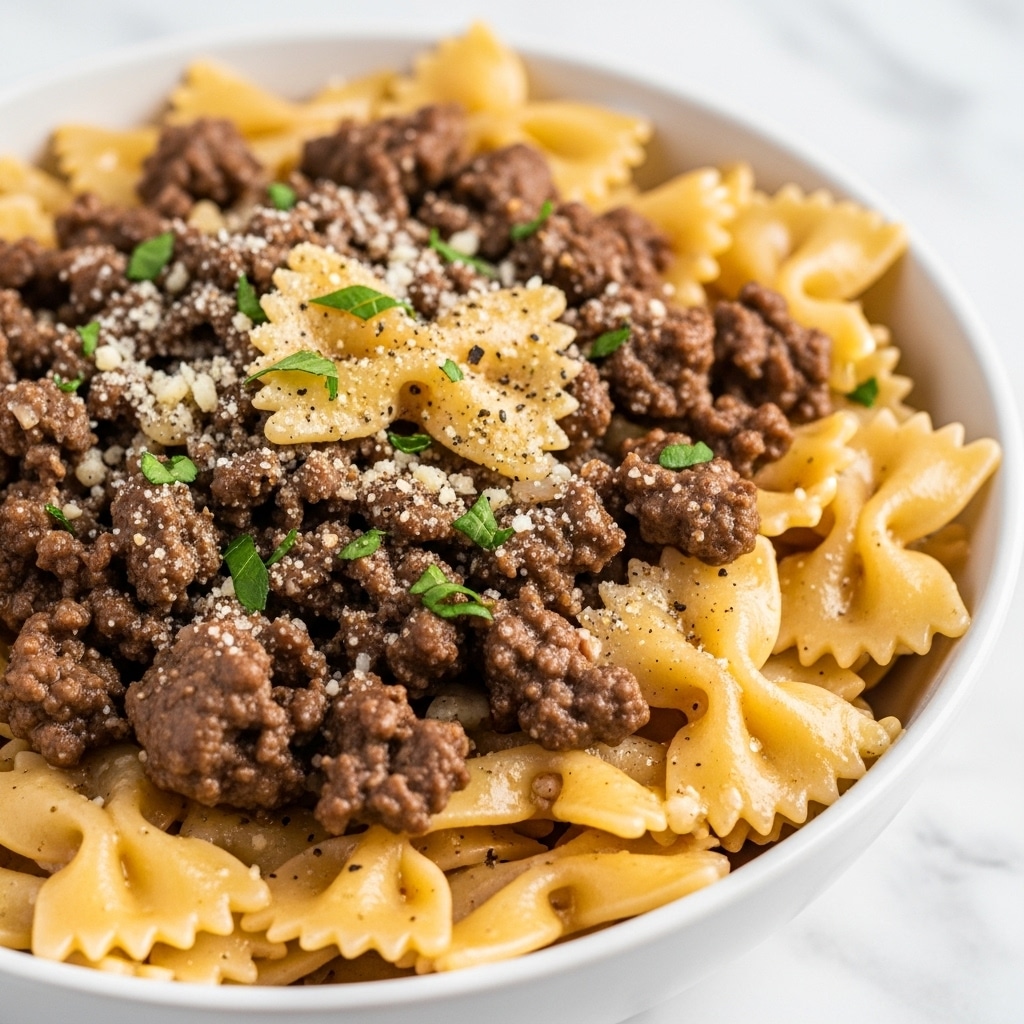A close-up view of a white bowl filled with farfalle pasta mixed with browned ground meat. The pasta is golden-colored with a slight shine from the sauce. The meat pieces are dark brown and crumbly, spread evenly throughout the pasta. Small green herb bits are sprinkled on top along with a light dusting of grated cheese and black pepper. The background shows a soft, white marbled texture. photo taken with an iphone --ar 4:5 --v 7