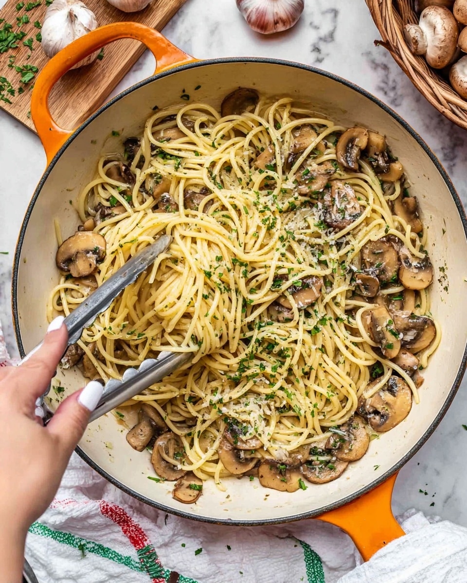 A large white pan with orange handles holds a dish of spaghetti mixed with sautéed sliced mushrooms, sprinkled lightly with chopped green herbs and ground black pepper. The spaghetti strands are pale yellow and thin, twisting through the brown and tender mushrooms. A woman's hand with white nail polish is using silver tongs to lift some spaghetti from the pan. The pan sits on a white marbled surface with a white cloth underneath, decorated with green and red lines. Nearby are a wooden board with herbs, a basket of garlic, and some whole mushrooms. photo taken with an iphone --ar 4:5 --v 7