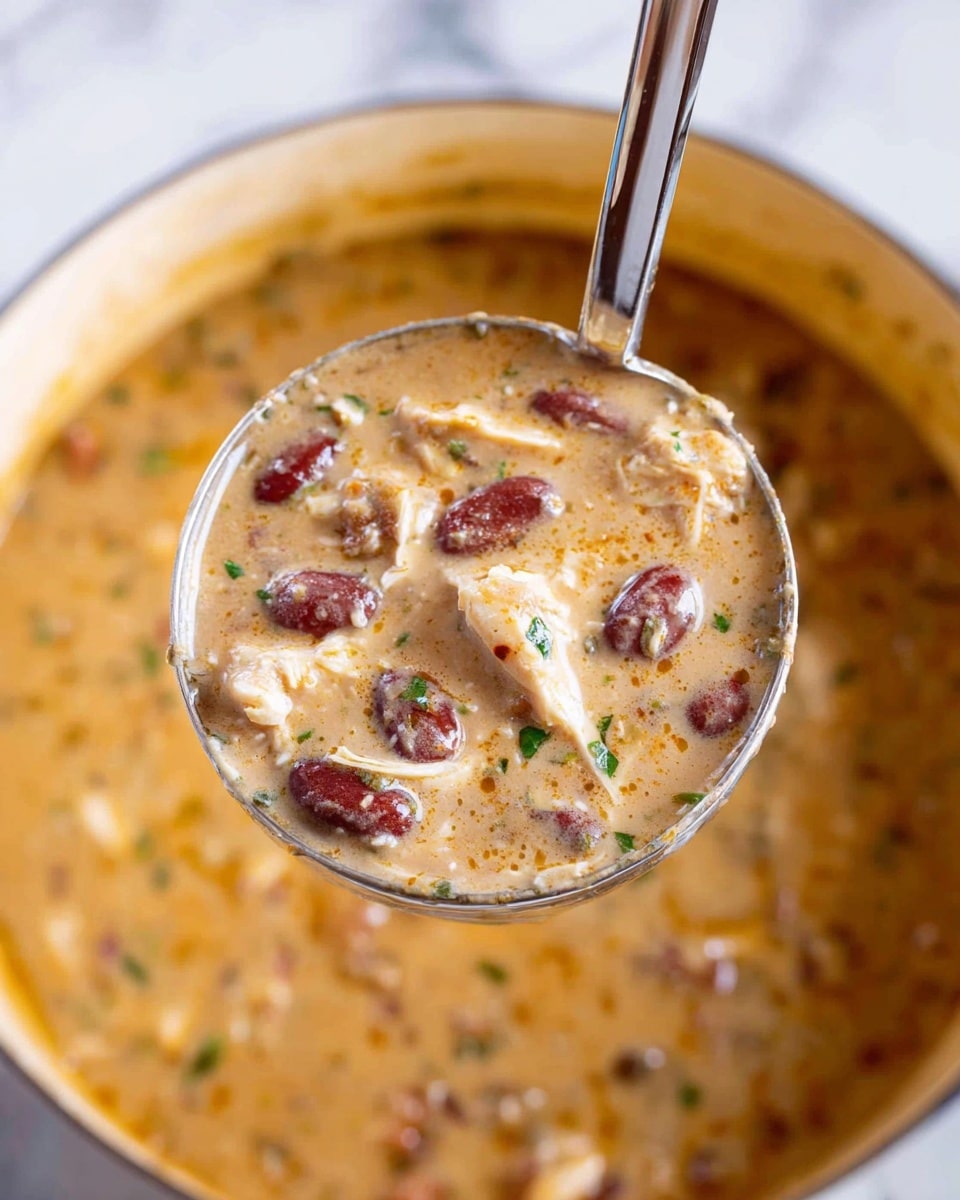 The image shows a close-up of a creamy chili soup in a white bowl, with three main layers visible: a base layer of thick orange broth with a smooth texture, a middle layer filled with red kidney beans and pieces of corn kernels scattered evenly, and a top layer of shredded cooked chicken and brownish ground meat chunks, all sprinkled with finely chopped green herbs. The dish looks rich and hearty, placed on a white marbled surface with some green leaves and a silver spoon blurred in the background. Photo taken with an iphone --ar 4:5 --v 7