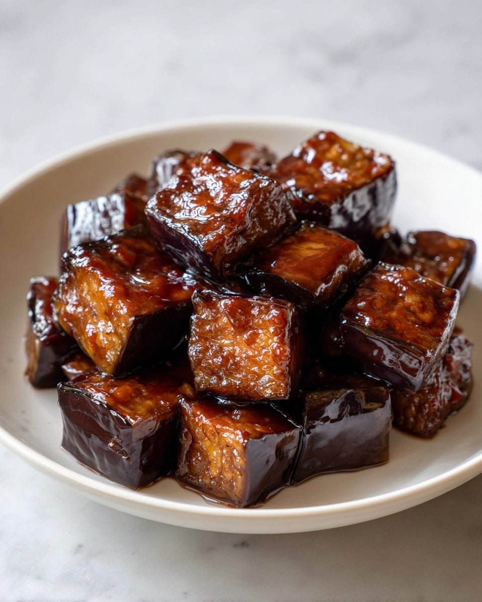 A close-up view of a heap of glossy, dark brown cubes of cooked eggplant coated in a shiny rich sauce, sitting in a white bowl. Each cube shows a contrast between the deeper brown edges and the lighter, slightly textured interior. The sauce adds a wet, sticky gloss that highlights the smooth, slightly wrinkled skin of the eggplant. The bowl rests on a white marbled surface with soft natural light giving the dish a warm, inviting look. photo taken with an iphone --ar 4:5 --v 7