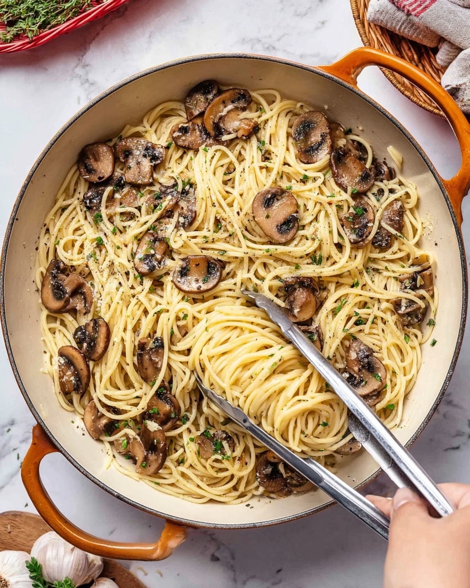A large white pan with orange handles is filled with a single layer of cooked spaghetti, light yellow in color and slightly glossy. Mixed evenly on top are sautéed mushroom slices, brown with a soft texture and a few darker charred edges. The pasta and mushrooms are sprinkled with small bits of green herbs and black pepper, adding tiny specks of green and black across the dish. A woman’s hand holding shiny silver tongs is lifting some of the spaghetti with mushrooms. The pan sits on a white marbled surface with a small wooden board holding fresh herbs and a basket with garlic bulbs nearby. photo taken with an iphone --ar 4:5 --v 7