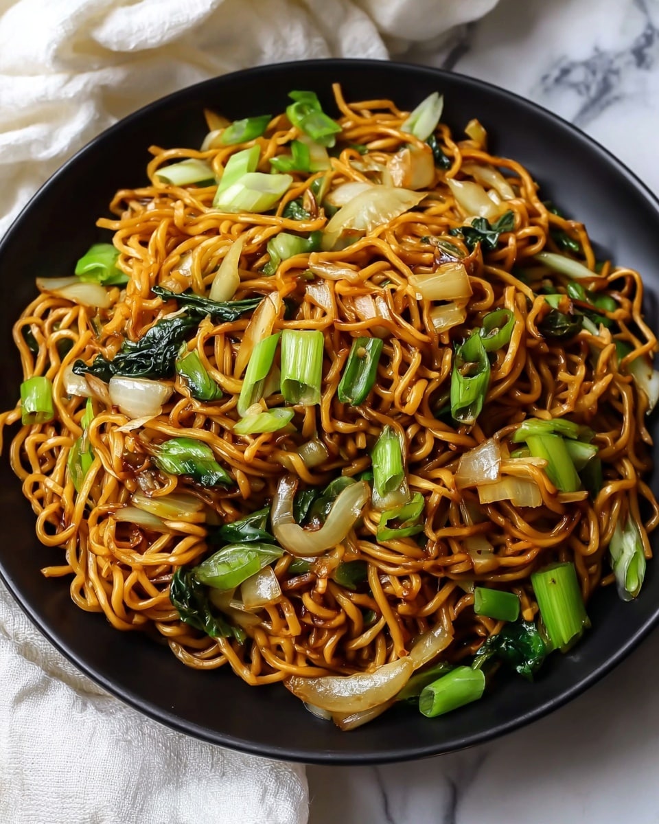 A black bowl filled with one main layer of golden-brown stir-fried noodles mixed with bright green chopped spring onions, translucent cooked white onion pieces, and small bits of dark green leafy vegetables, all slightly glossy from a savory sauce. The noodles look soft and slightly twisted, spread evenly across the bowl. The bowl is placed on a white marbled surface with a white fabric cloth partly visible around it. photo taken with an iphone --ar 4:5 --v 7