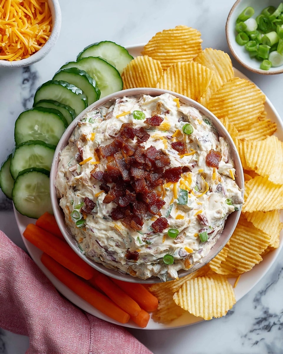 A bowl filled with a thick and creamy dip showing visible bits of green onions and shredded orange cheese mixed throughout, topped with small pieces of crispy dark red bacon. Surrounding the bowl on a white plate are fresh cucumber slices with dark green edges, bright orange carrot sticks, and yellow ridged potato chips. A small bowl with sliced green onions and a pink cloth napkin are nearby on a white marbled surface. photo taken with an iphone --ar 4:5 --v 7