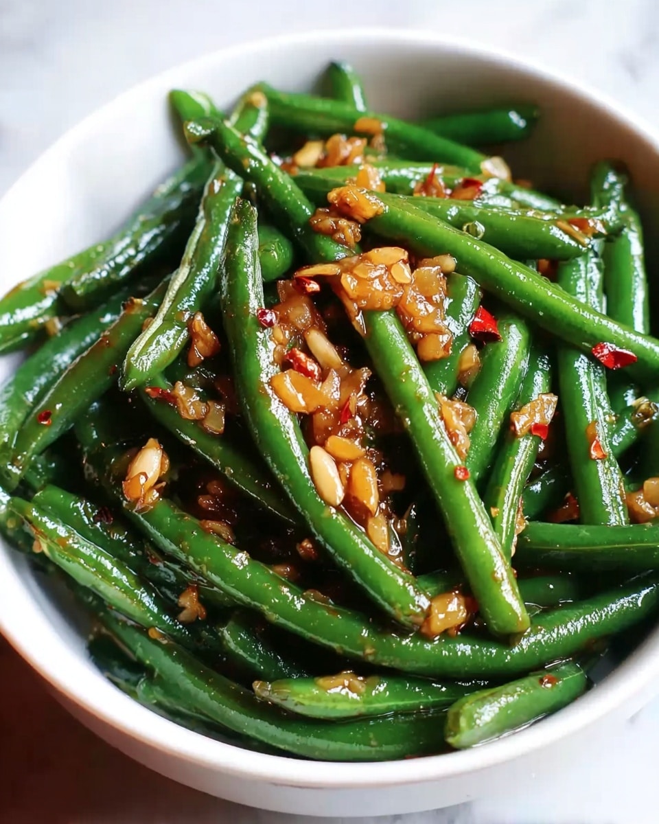 A close-up view of a bowl filled with cooked green beans coated in a glossy sauce, topped generously with pieces of golden-brown sautéed garlic and small red chili flakes scattered throughout; the green beans look tender and shiny, slightly wrinkled from cooking, and the bowl is white, sitting on a white marbled surface. photo taken with an iphone --ar 4:5 --v 7