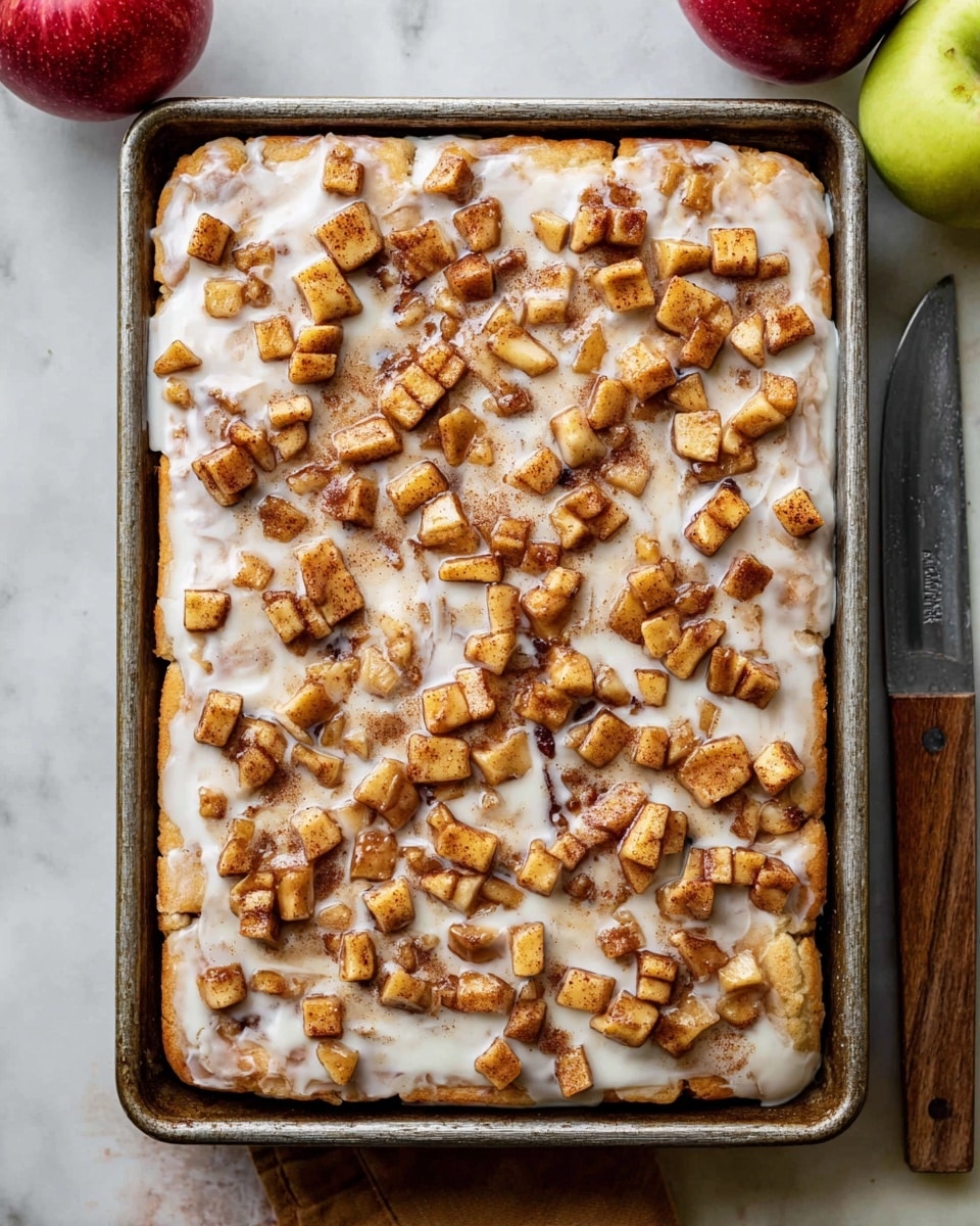 A rectangular cinnamon roll cake in a metal baking pan sits on a white marbled surface with a red apple on the left and a green apple on the right. The cake has two visible layers: a golden-brown baked dough base and a thick white glaze layer spread on top. Scattered all over the glaze are small cubes of caramelized cinnamon-spiced apples that add a textured brown and golden color. To the right of the pan, a knife with a wooden handle rests on the surface. photo taken with an iphone --ar 4:5 --v 7