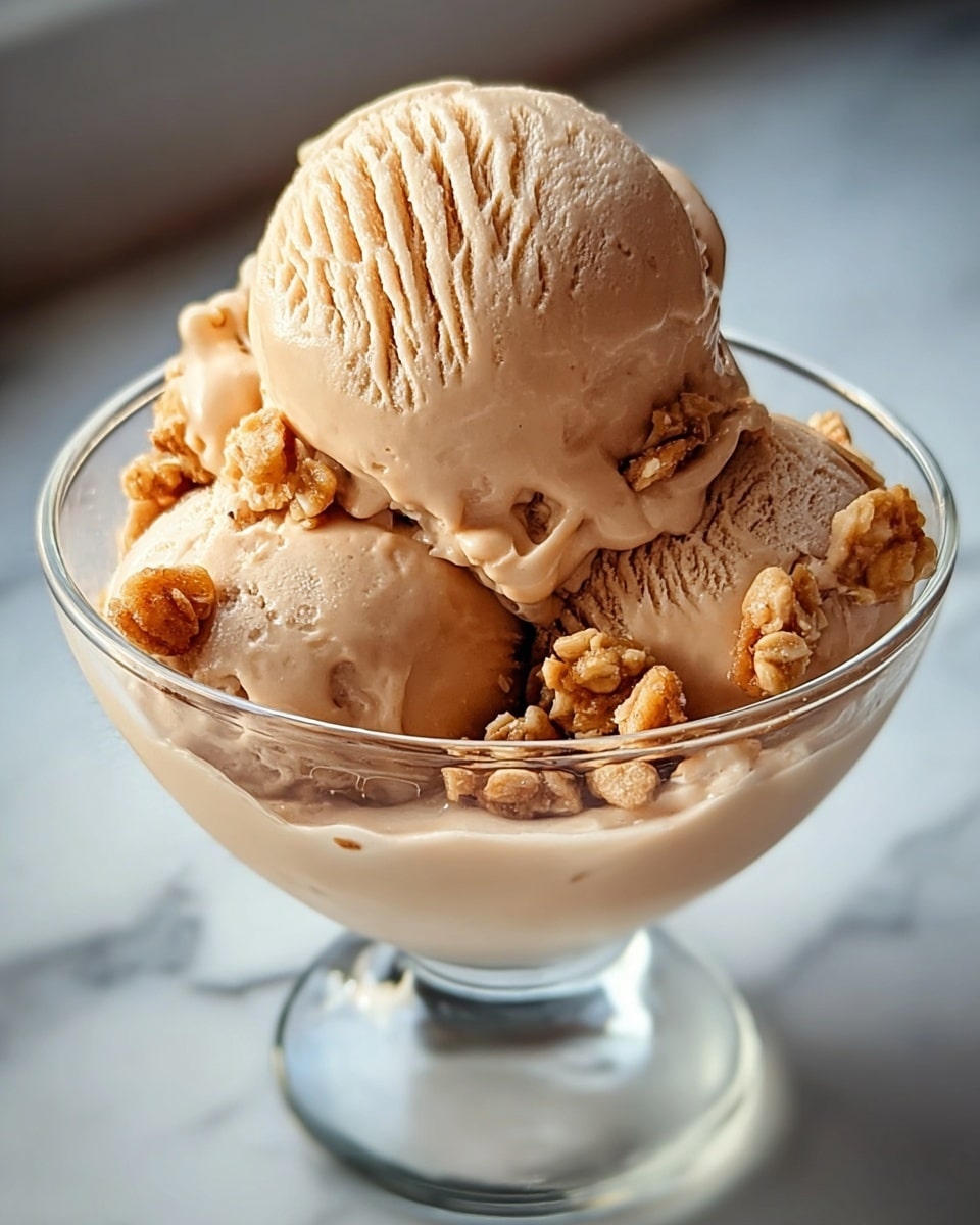 A close-up view of a glass dessert cup filled with three smooth, light brown ice cream scoops, slightly shiny and creamy in texture. The top scoop is larger and shows visible ridges from scooping, sitting above two smaller scoops that are nestled closely. Scattered around the ice cream scoops are small clusters of light golden granola adding a crunchy texture contrast. The cup is placed on a surface with a white marbled texture, and soft natural light highlights the creamy and crunchy layers. photo taken with an iphone --ar 4:5 --v 7