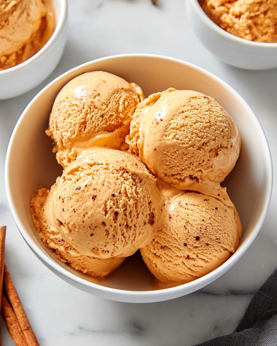 A white bowl filled with four round scoops of smooth, creamy pumpkin-colored ice cream that has small specks of spices visible, giving it a textured look; the ice cream appears slightly melting with a glossy finish. The bowl is placed on a white marbled surface, with cinnamon sticks nearby and parts of two other white bowls with similar ice cream scoops visible in the background. The lighting highlights the soft texture and warm color of the ice cream, creating a cozy feel. photo taken with an iphone --ar 4:5 --v 7