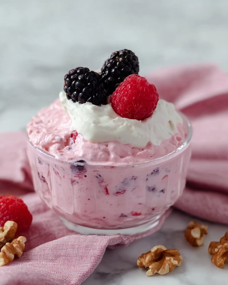 A clear glass bowl filled with a fluffy pink mousse that has small dark berry bits mixed inside, topped with a dollop of smooth white cream. On top of the cream, there are two large shiny black blackberries and one bright red raspberry, along with a few small pieces of light brown walnuts scattered around. The bowl rests on a soft pink and white cloth over a white marbled surface. Photo taken with an iphone --ar 4:5 --v 7