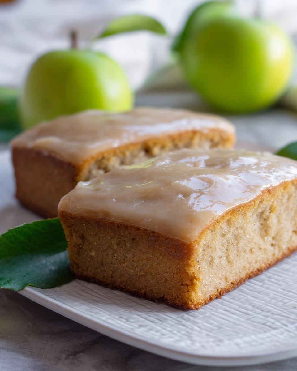 The image shows a close-up of two square pieces of moist cake with a smooth, light tan glaze covering the top layer. The cake has a soft texture with a slightly browned edge. In the background, there are a few small green apples with leaves, adding a fresh touch. The pieces are placed on a white plate that has a subtle textured design, and the setup sits on a white marbled surface. The overall scene is bright and natural. photo taken with an iphone --ar 4:5 --v 7