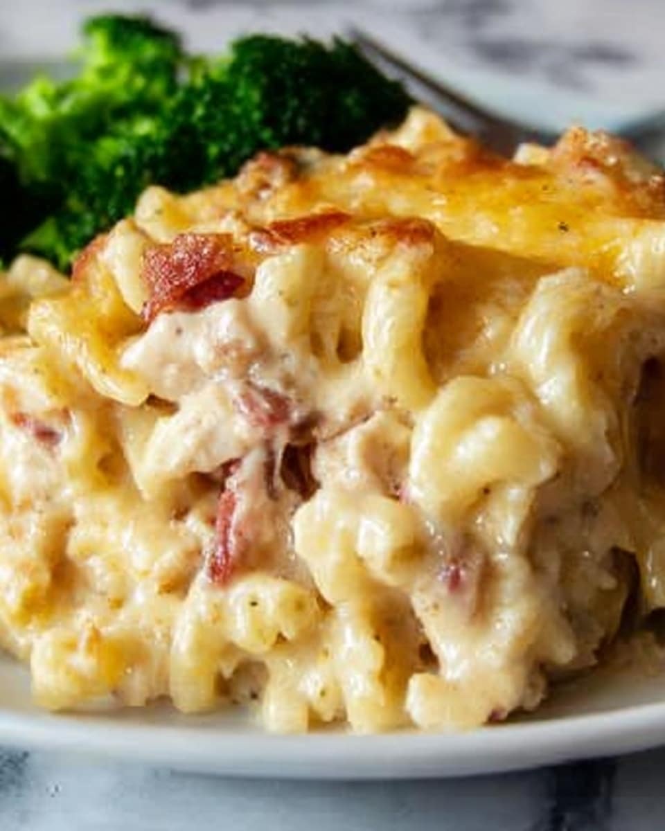 The image shows a close-up of a creamy macaroni and cheese dish with three clear layers: the top layer is melted, slightly browned cheese with a smooth, gooey texture; underneath is a thick, pale yellow cheese sauce that coats the curly pasta loved with chunks of pinkish cooked chicken and bits of reddish bacon; the bottom layer is the white curly pasta that is soft and slightly firm. The dish is served on a white plate with a side of green broccoli, all placed on a white marbled surface. Photo taken with an iphone --ar 4:5 --v 7